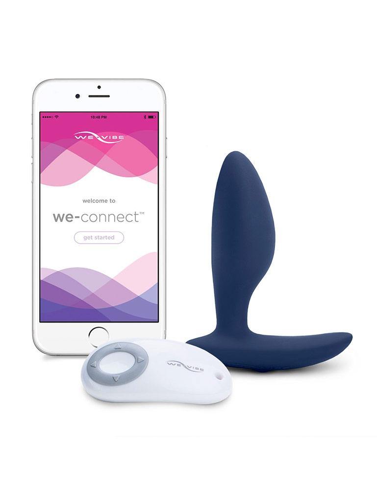 The We-Vibe Ditto Vibrating Butt Plug is shown next to its remote and a white iPhone against a blank background. The remote is oval-shaped with its grey buttons arranged in a circle. The iPhone displays the homepage of the WeVibe Connect app.
