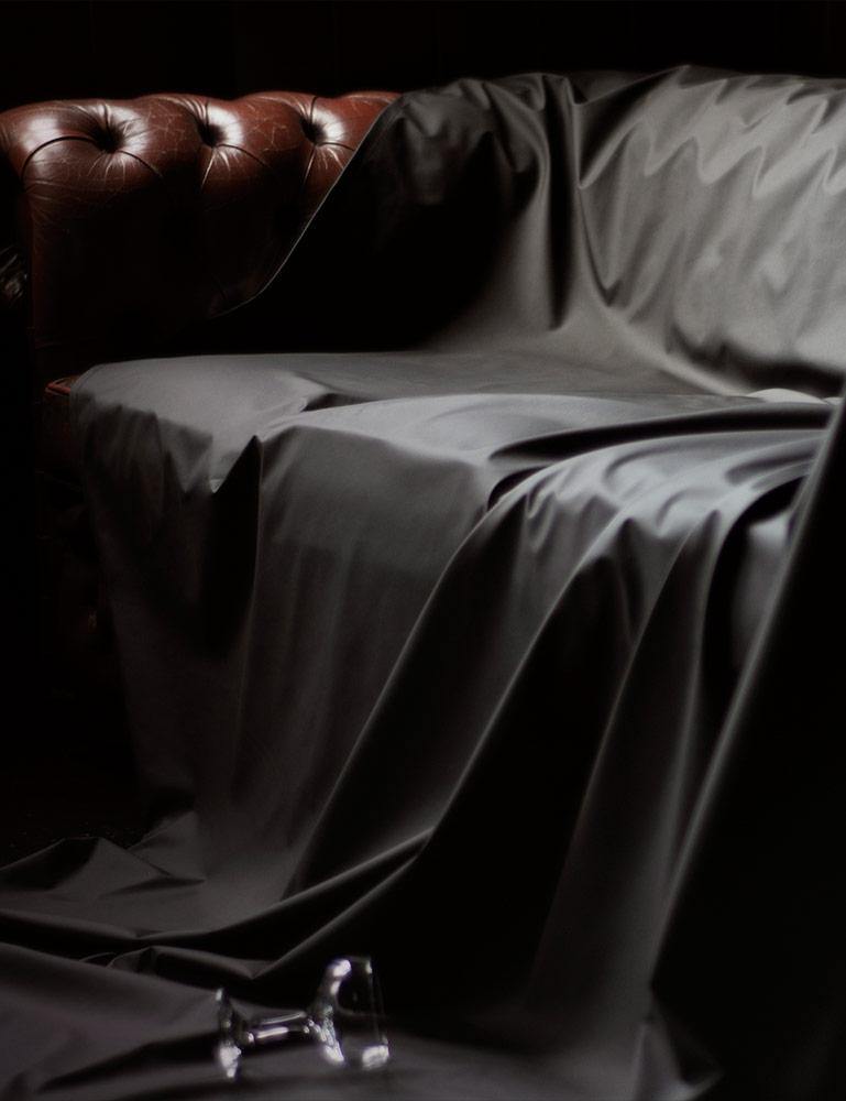 A brown leather couch is shown against a black background. All but the arm of the couch is covered by the Fluid Proof Protective Throw By Sheets Of San Francisco, a black sheet that drapes onto the floor. On the floor is a knocked-over glass on the sheet.