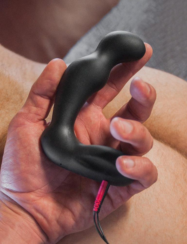A man’s hand rests on somebody’s ass, holding the Electrastim "Sirius" Silicone Noir Prostate Massager, made of black silicone. The plug has a widened mid-section and a bulbous tip. The base is wide and extended on one side, with 2 black wires plugged into it.
