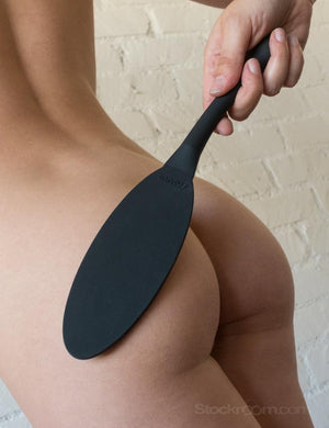 A close-up of a woman’s bare butt and back is shown in front of a white wall. Somebody is holding the black Tantus Gen Mini Silicone Paddle against her butt.