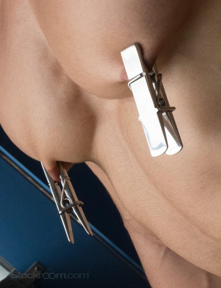A closeup of a nude man’s chest is shown against a dark blue background. He has the Stainless Steel Clothespin Nipple Clamps on his nipples.