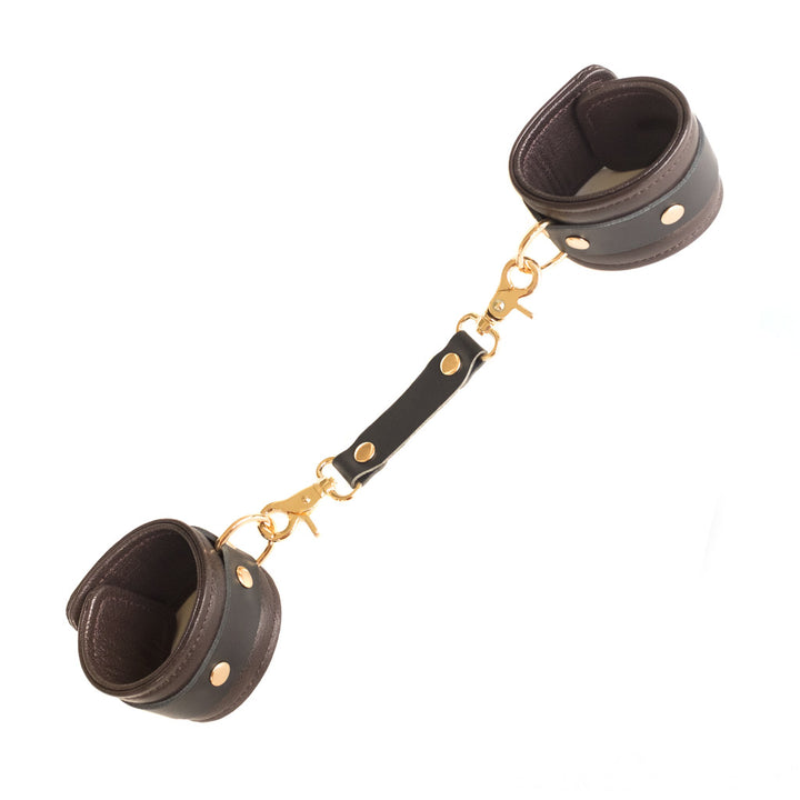 The Brown Leather Wrist Restraints With Gold Accent Hardware are shown against a blank background. They are made of a wide piece of leather with a smaller, darker piece riveted around it. A matching restraint clip connects the cuffs by their D-rings.
