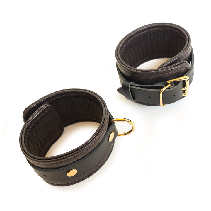 The Brown Leather Ankle Restraints With Gold Accent Hardware are shown against a blank background. They are made of a wide piece of leather with a smaller, darker piece riveted around it.