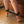 Load image into Gallery viewer, A close-up of a person’s lower legs and bare feet is shown. Their feet are pointed with their toes touching the wood floor, and they are wearing the Brown Leather Ankle Restraints with Gold Accent Hardware, which are clipped together with the included restraint clip.
