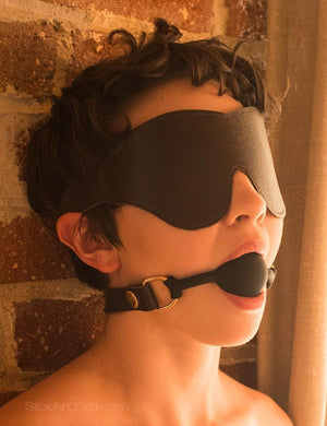 A close-up of a woman with short brown hair is shown with her head leaning back against a brick wall. She has a black ball gag in her mouth and wears the Classic Cut Brown Leather Blindfold. 