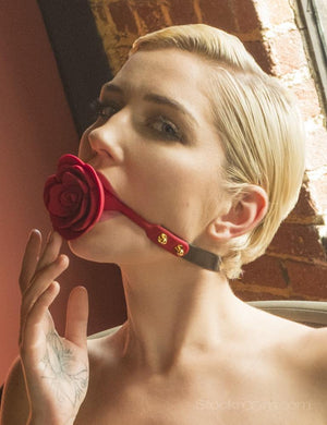 A close-up of a woman with short blonde hair sitting in front of a brick wall with her hand raised to her mouth. In her mouth is the Rose Ball Gag.