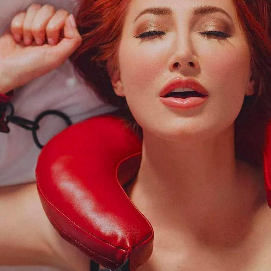 A woman with red hair is shown laying on a bed from the shoulder up. Her neck is positioned in the Saffron Thigh Sling, and her wrists are cuffed to the sling. She has an expression of pleasure on her face.