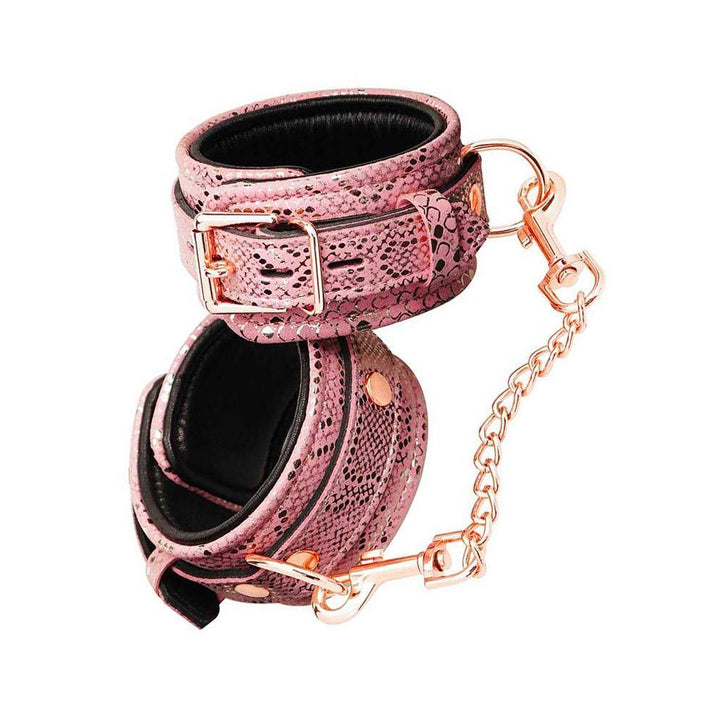 A pair of Rose Gold Snake Wrist Cuffs are shown against a blank background. The cuffs have a pink snakeskin design and rose gold hardware, including a lockable buckle, D-rings, and an included chain with claw hooks at the ends. 