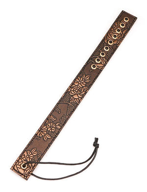 The Vegan Floral Ruler With Gems is shown against a blank background. It is a piece of brown faux leather shaped like a ruler with a floral design in light brown. There is a row of black gems at the top of the ruler and a wrist loop at the bottom.