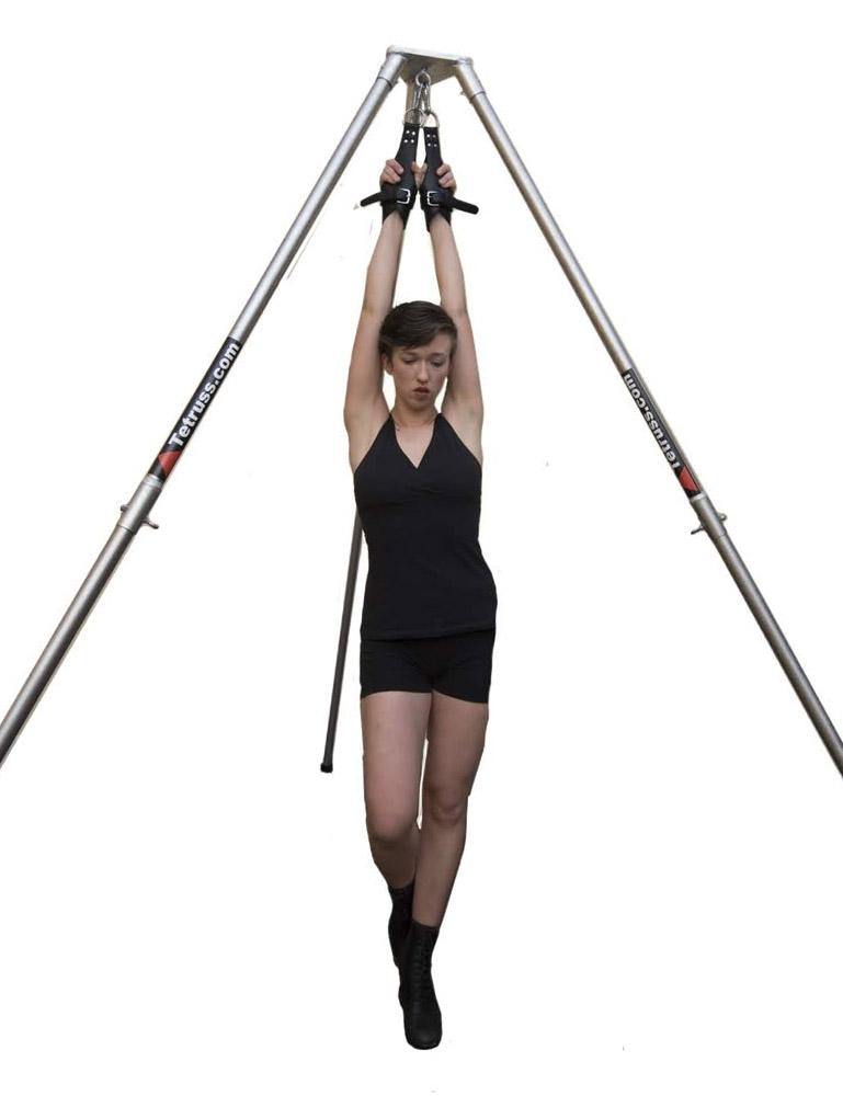The Tetruss Suspension Bondage Frame, made of 3 silver poles in a triangle formation, is shown in use. A woman in a black tanktop and shorts is wearing black suspension cuffs, which are chained to the top of the frame, lifting her arms above her head.