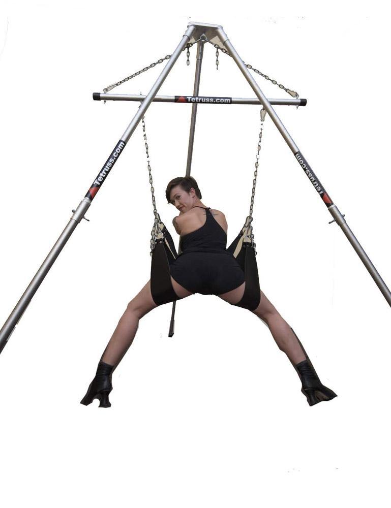  The Tetruss Portable Dungeon Deluxe Bundle is shown in use against a blank background. A woman is shown from the back, leaning forward against one of the stirrups while her legs are supported by the others, all of which are attached to the spreader bar.