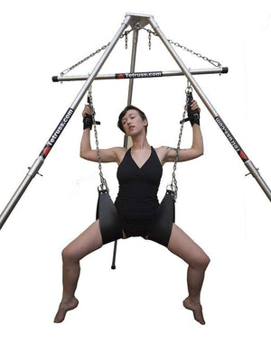 The Tetruss Portable Dungeon Deluxe Bundle is shown in use against a blank background. The spreader bar is connected to the frame, supporting a woman sitting in the leather slings. Her suspension wrist cuffs are attached to the spreader bar.
