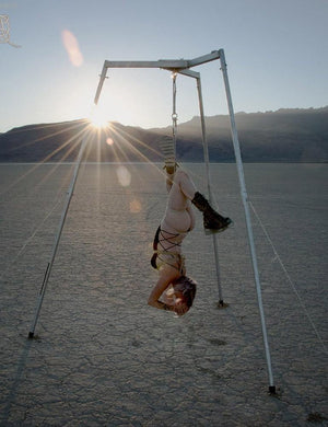 A woman is shown suspended from the Tetruss Maxximus Suspension Bondage Frame. She is outdoors in the desert and is hanging upside down, suspended by ropes tied around her torso.