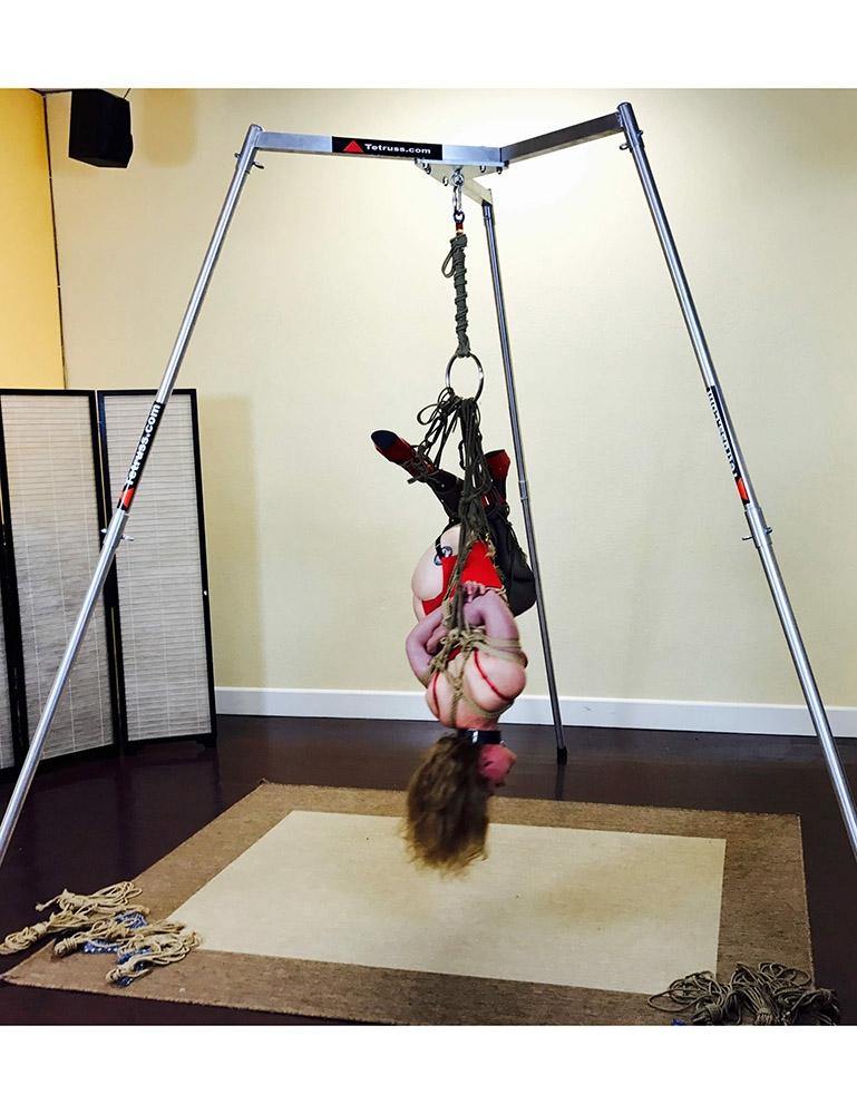 A woman is suspended from the Tetruss Maxximus Suspension Bondage Frame. Her arms are tied behind her back, and she is suspended upside down by them. She is wearing a red bodysuit and long black boots. Her face is visibly red. 