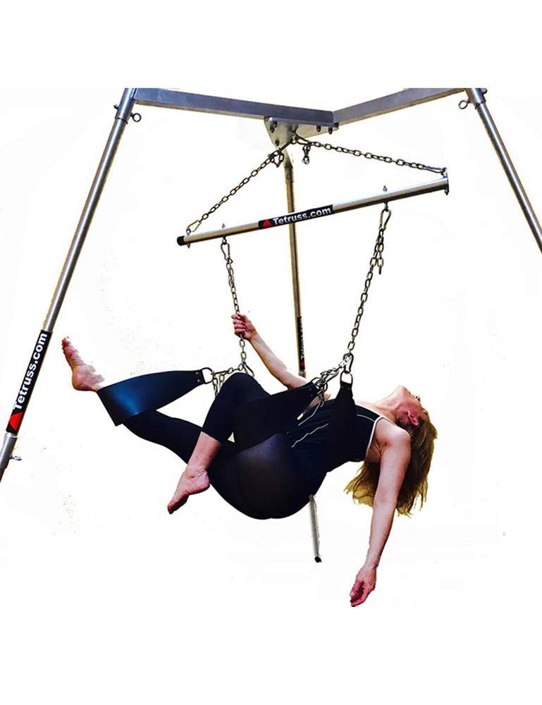 The Tetruss Maxximus Portable Dungeon Deluxe Bundle is shown in use against a blank background. The metal bondage frame has a spreader bar attached to the center, which supports 3 leather stirrups. A woman in a black bodysuit is reclining in the stirrups.