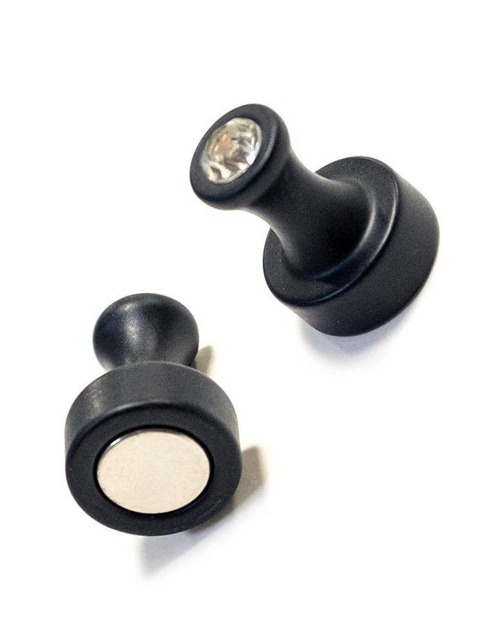 A pair of Magnetic Nipple Power Pinchers are displayed against a blank background. They are black and resemble the back of a thumbtack with a small crystal at the top. There is a magnet at the base of the pinchers.