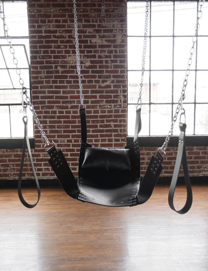 The black Leather Sling with metal rivets is suspended from the ceiling by chains in a brick room. It has a large central piece and two smaller leather pieces for suspension on top and bottom. Stirrups hang from the chains at the bottom of the swing.