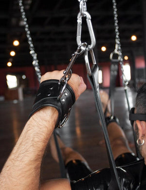 A close-up of a man’s wrist, wearing black leather wrist cuffs, is shown clipped to the hardware suspending the Leather Sling with a metal snap hook.