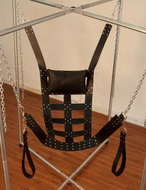 The black Leather Web Style Swing is shown suspended from a metal frame. The Sling is made of woven pieces of black leather secured together with metal rivets. It has leather thigh stirrups on each side.