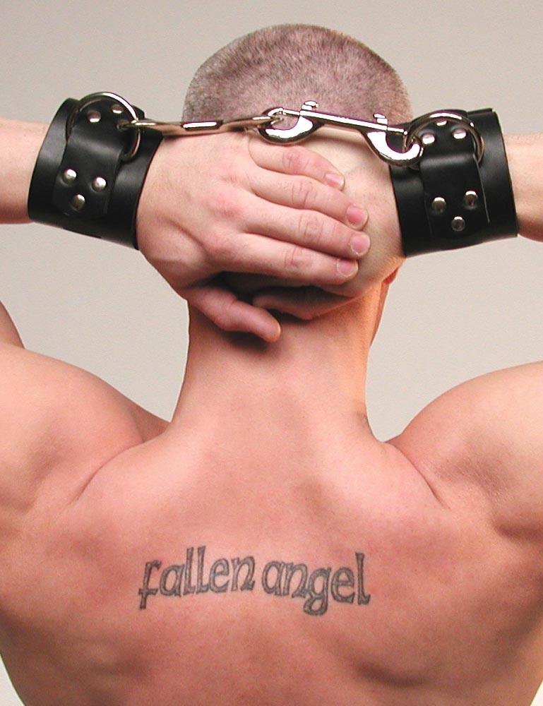 A muscular man is shown from behind with his hands clasped behind his head. He wears the Extra Wide Black Leather BDSM Wrist Cuffs with metal hardware. The cuffs are connected to each other with two snap hooks attached to the D-rings on the cuffs.