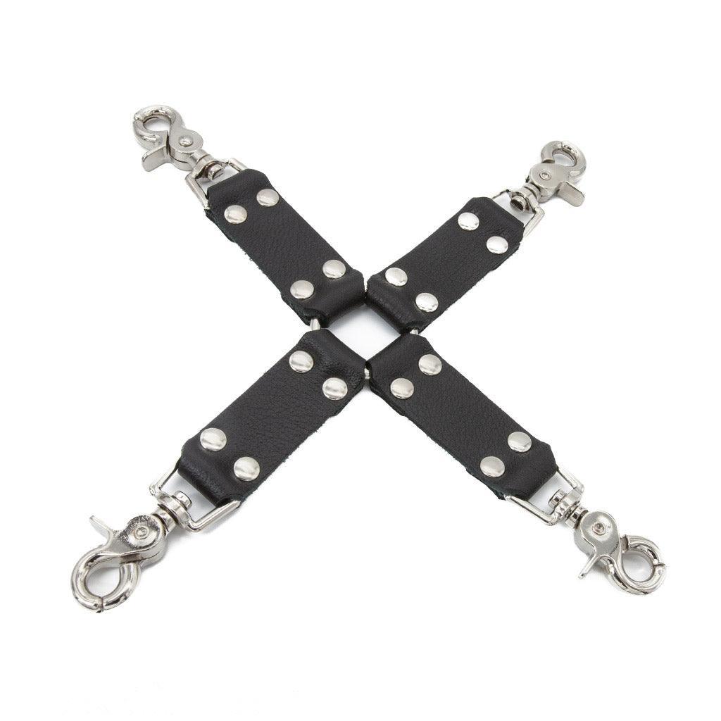 The black Leather Hog Tie is displayed against a blank background. A metal O-ring is in the center with four leather strips attached to it in the shape of an X. Each leather strip has a metal snap hook on the end.