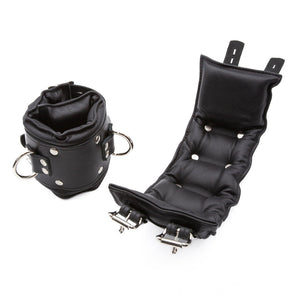 The Premium Lockable Dungeon BDSM Leather Ankle Cuffs are shown against a blank background. They are made of black leather with silver hardware. The cuffs are wide and padded. They have a D-ring and two adjustable straps with lockable buckles.