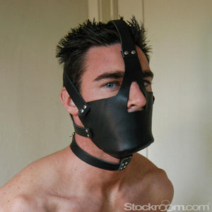 A shirtless man with dark, spiky hair is shown from the shoulders up in front of a beige wall. He wears the Leather Head Harness with Muzzle. It is a muzzle made of black leather which covers his mouth and chin and straps around his head.