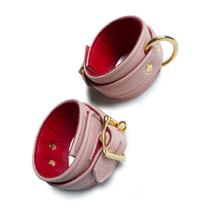 A set of Premium Garment Leather Ankle Cuffs With 18k Gold Plated Hardware are displayed against a blank leather background. The cuffs are made of light pink leather with a red inner lining. They each have one D-ring and fasten with a lockable buckle.