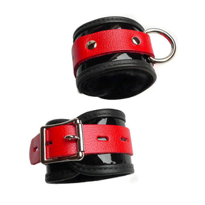 The Firecracker Patent Leather BDSM Wrist Restraints are shown against a blank background. They are black patent leather with a thin strip of red leather wrapped around them. They have a metal D-ring and lockable metal buckle.
