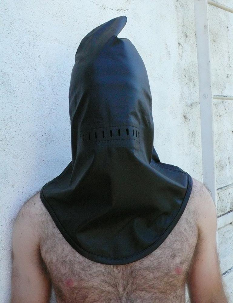 The Leather Guillotine Hood is shown on a man with dark chest hair standing in front of a white wall. The hood fits loosely and has a point at the top and a U-shaped bottom that reaches his chest.