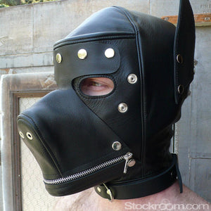 A close-up of a man wearing the Leather Dog Hood with Snap-on Muzzle, Blindfold and Gag and standing outdoors is shown. The hood covers his entire face, but the blindfold is removed. It has a muzzle that zips closed, puppy ears, and a buckled collar.