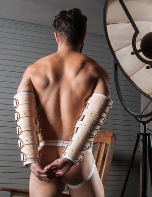 A muscular man in a jockstrap is shown from behind. His arms are in the BDSM Deluxe Medical Leather Arm Splints. The splints are made of tan leather with a white border and cover his wrist to his underarms. 