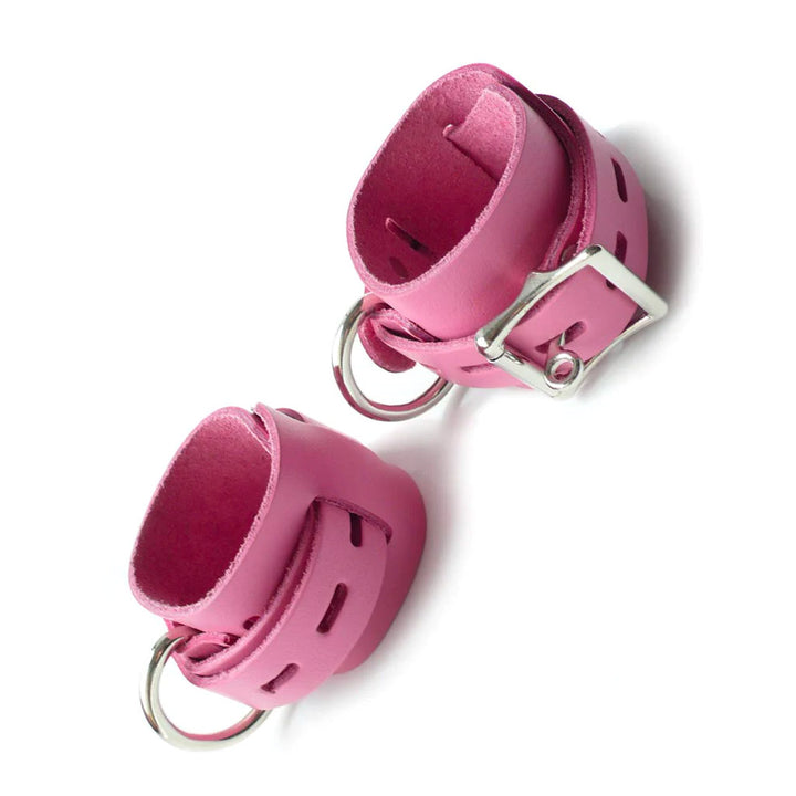 The Locking/Buckling Leather Cuffs are shown against a blank background. They are made of a wide piece of pink leather with a thinner, notched strip wrapping around them. They each have a metal D-ring and a lockable buckle.
