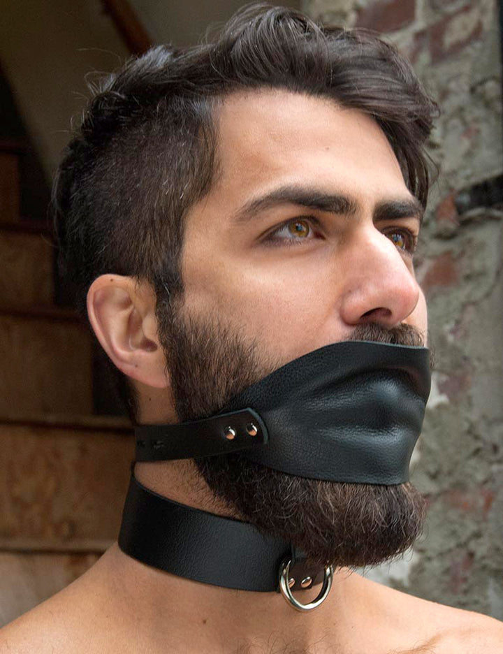 A dark-haired man’s face is shown in front of a stone wall. He has facial hair and is wearing the Over The Mouth Gag With A Silicone Ball and a black collar. The gag has a piece of black leather that completely covers his and a leather strap.
