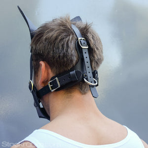 A man’s head is shown from behind. He is wearing the K9 Muzzle With Removable Silicone Ball Gag. It has one strap that comes over the back of his head vertically, and 2 that go across horizontally. All of the straps fasten with a silver buckle.