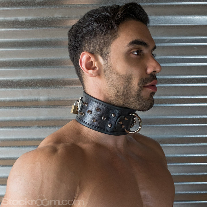 A close-up of a brunette man with facial hair is shown in profile. He wears the BDSM Alpha Dog Leather Collar with Spikes. The collar is wide and has a metal D-ring in the center and spikes covering it. It is padlocked in the back.