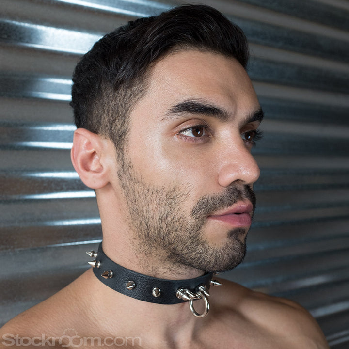 A close-up of a brunette man with facial hair shows him standing in front of a metal wall. He wears the black Leather Collar with Spikes, which has a dangling O-ring in the front and has evenly space conical spikes going around the collar.