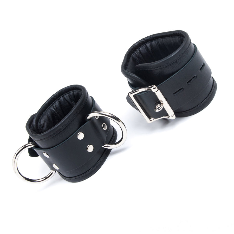The black leather Deluxe Padded Leather Wrist Restraints with D-Rings are shown against a blank background. The cuffs have silver buckles and two D-rings.