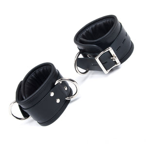 The Deluxe Padded Leather Ankle Restraints With D-Rings are shown cuffed against a blank background. They are made of black leather and have two silver D-rings and a silver buckle.