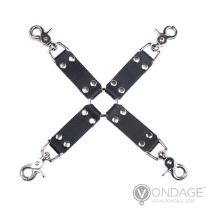 The Vondage Hog Tie is displayed against a blank background. It has a silver O-ring in the center with four pieces of black faux leather attached in the shape of an “X.” The leather pieces each have a metal snap hook at the end.