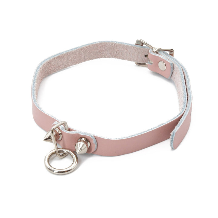 The Stupid Cute choker is shown against a blank background. It is a light pink leather choker with metal hardware, including a dangling O-ring and two spikes. It has an adjustable strap and buckles in the back.