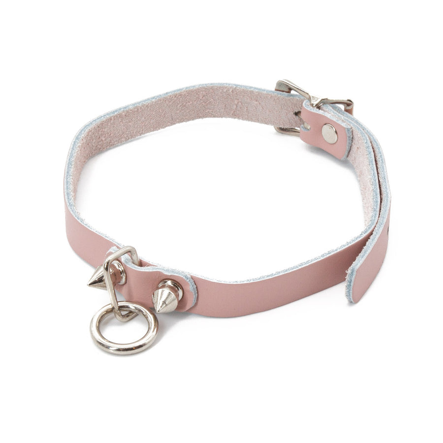The Stupid Cute choker is shown against a blank background. It is a light pink leather choker with metal hardware, including a dangling O-ring and two spikes. It has an adjustable strap and buckles in the back.