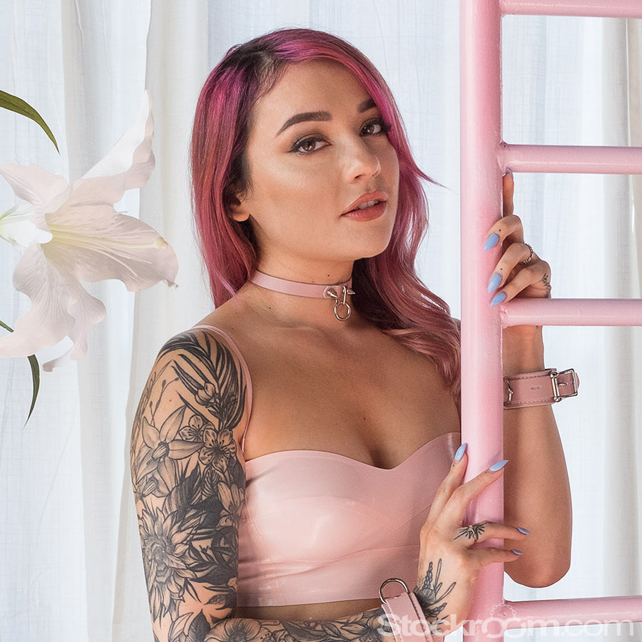 A woman with pink hair wearing a pink latex dress poses next to a pink metal structure. She wears the baby pink Stupid Cute Choker and matching Stupid Cute wrist cuffs.