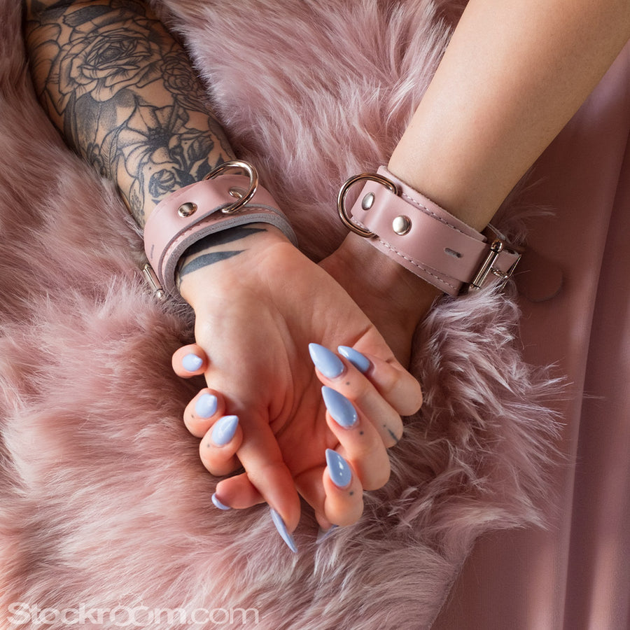 A close-up of a woman’s hands with pointed lavender nails are shown resting on top of each other on a furry pink blanket. She is wearing the Stupid Cute Wrist Cuffs, which are made of light pink leather.