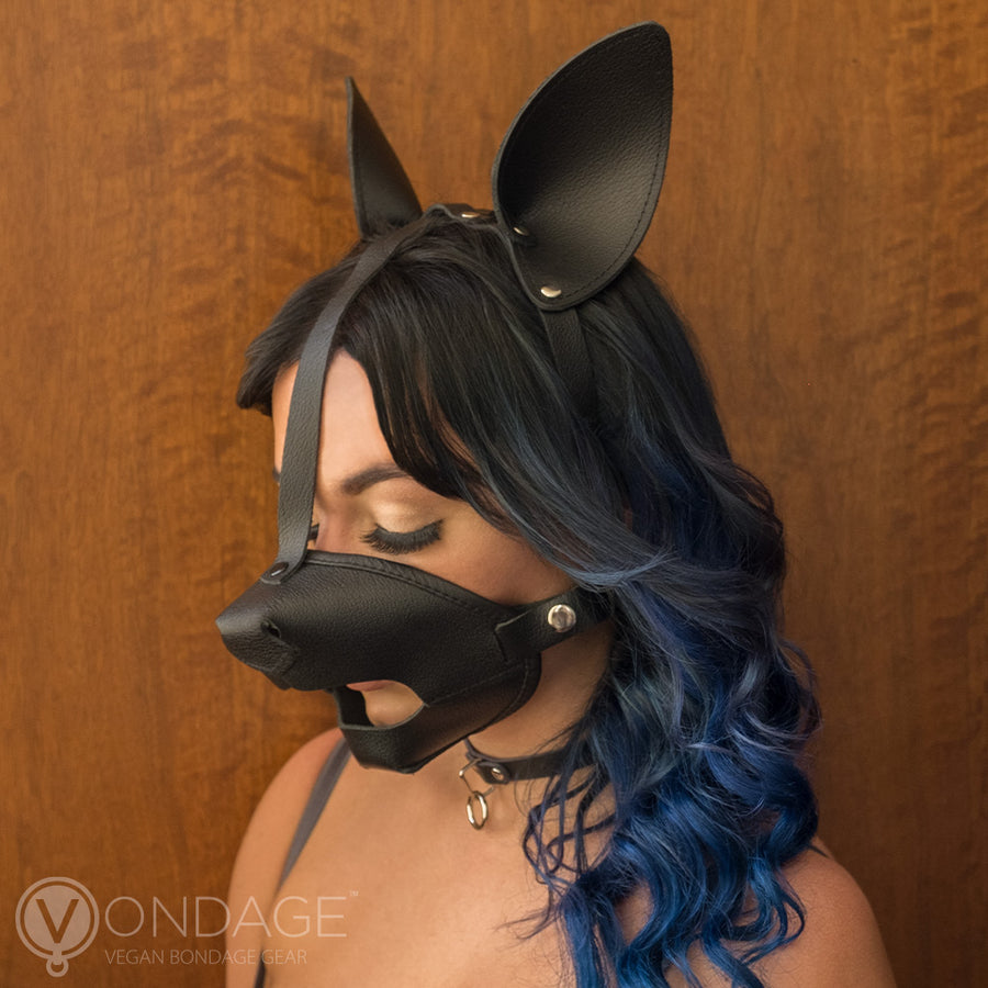 A woman stands against a wall with her eyes lowered. She wears the black Vondage Pet Play K9 Muzzle with Removable Ball Gag. The muzzle is shaped like a dog snout and covers her lower face, but has a mouth opening. It connects to black dog ears.