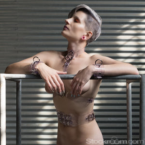 A woman with short silver hair leans on a metal structure, looking upwards. She wears the Clear CTRL Wrist Cuffs, as well as a matching waist cincher and bust harness.