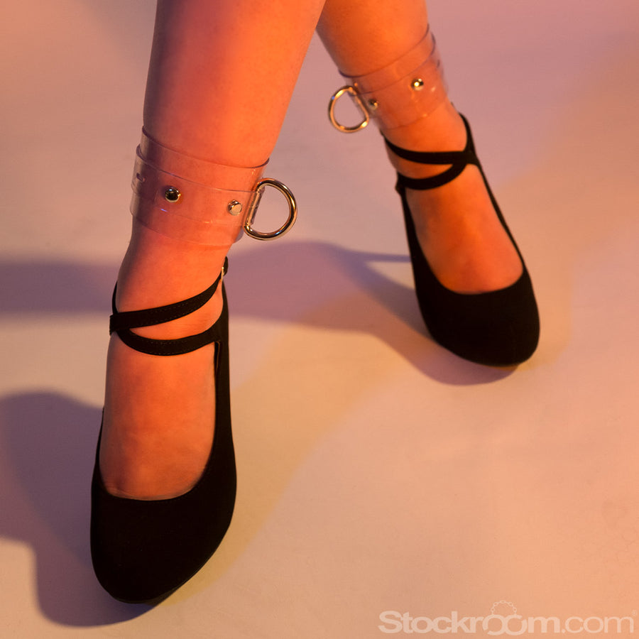 A close-up of a person's feet in black heels with straps wrapped around their ankles is shown in pinkish-purple lighting. They wear the Clear CTRL Vinyl Ankle Cuffs, which have a thick band of transparent PVC and a D-ring on each cuff.