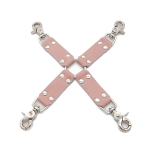 The Stupid Cute Hog Tie is shown against a blank background. It is made of four pink leather strips in the shape of an 'X,' attached in the center by a metal O-ring. Each leather strap has a metal snap hook at the end.