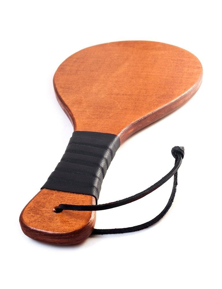 The Leather Wrapped Round Wood Spanking Paddle is shown against a blank background. The paddle is made of light-colored wood and has a black wrist loop at the bottom. The middle of the handle is wrapped in a black leather strip.