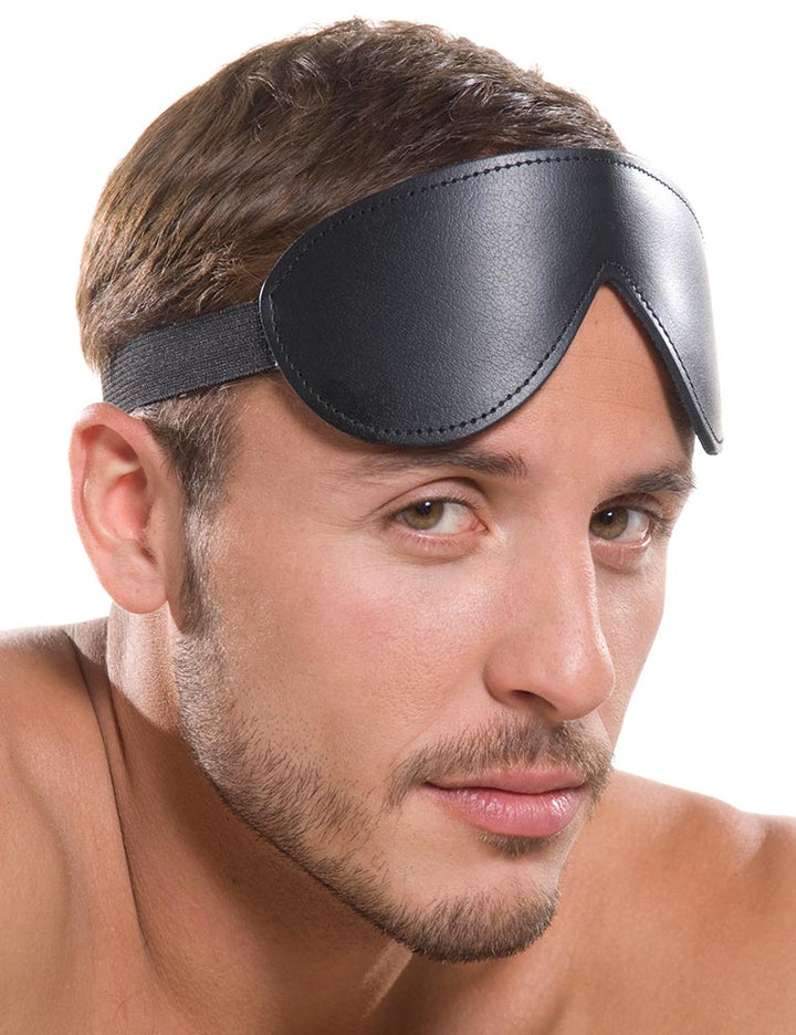 A close-up of a brunette man with light facial hair is shown against a blank background. He has the black Kinklab Bondage Basics Padded Leather Blindfold pulled above his eyes.